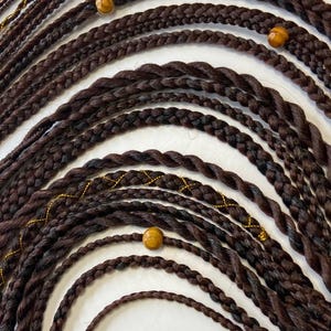 May include: Close-up of dark brown braided hair extensions with wooden beads and gold accents. The braids are arranged in a curved pattern, showcasing the texture and detail of the hair. The overall aesthetic is natural and stylish.