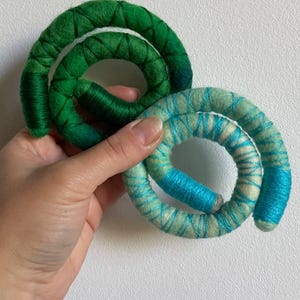 May include: Two spiral-shaped decorative objects. One is green, the other is blue and white. The objects are made of a soft, textured material, possibly yarn or felt. The spirals are held in a hand against a white background.