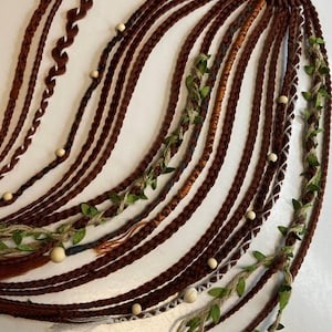 Può includere: Extension per capelli intrecciati marroni con elementi decorativi. Le extension sono attaccate a un elastico nero. Alcune trecce sono decorate con perline di legno e foglie verdi.