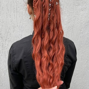 May include: A person's back is shown, with long, wavy, reddish-orange hair styled in braids and extensions. Silver-colored decorative accents are woven into the braids. The person is wearing a black shirt. A hand holds the hair.
