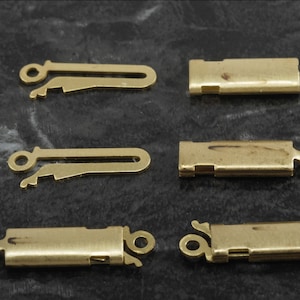 May include: Six gold-coloured jewellery clasps are shown against a dark background. The clasps are rectangular with a loop on one end and a hook-and-eye closure on the other. They are likely used for necklaces or bracelets.