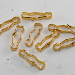 Puede incluir: Un grupo de diez cierres metálicos de color dorado, también conocidos como broches de presión, están esparcidos sobre una superficie blanca. Los cierres miden aproximadamente 2,5 cm de largo y tienen una forma curva.