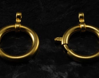 Op de afbeelding: Twee goudkleurige, cirkelvormige sieradssluitingen. Elke sluiting heeft een scharnierende ring met een kleine, bevestigde lus aan de bovenkant en een sluitmechanisme aan de zijkant. De sluitingen staan tegen een donkere achtergrond.