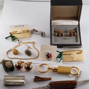 May include: Assortment of gold-toned jewelry and accessories including cufflinks, tie clips, a key chain, a pocket knife, and a pin. The items are displayed on a white surface with a few pieces in their original packaging.