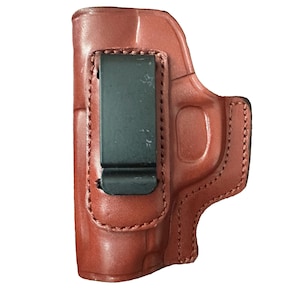 Pode incluir: Um coldre de couro castanho com um acabamento liso e moldado, apresentando um clipe de cinto de metal preto resistente fixado no exterior. O coldre tem costuras reforçadas ao longo das bordas e uma forma contornada para segurar uma arma.