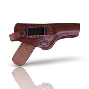 Op de afbeelding: Een bruine lederen holster met een gladde, gepolijste afwerking en zichtbare stiksels langs de randen. Het bevat een zwarte metalen riemclip aan de bovenkant en een leren riempje dat aan de zijkant is vastgezet met een donkere metalen drukknoop.