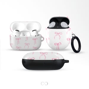 Puede incluir: Fundas protectoras blancas y negras para auriculares inalámbricos. Las fundas están decoradas con un patrón de lazos rosas. Una funda está abierta, revelando los auriculares. Un mosquetón negro está sujeto a una funda.