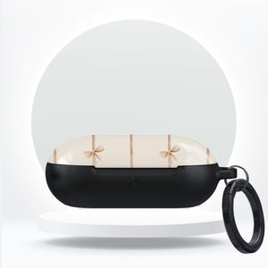 Könnte beinhalten: Schwarzes und cremefarbenes Kopfhörer-Etui mit dekorativem Schleifenmuster. Das Etui hat eine schwarze Basis und eine cremefarbene Oberseite mit vertikalen Streifen und kleinen Schleifenakzenten. Ein schwarzer Ring ist an der Seite befestigt.