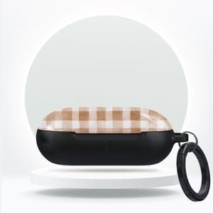 May include: A black protective case for wireless earbuds with a tan and white plaid pattern on the lid. The case has a black ring for attaching to a keychain or bag. The case is on a white pedestal.