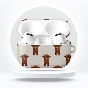 May include: A beige AirPods case featuring a repeating pattern of brown Highland cows. The case is open, revealing white AirPods inside. The case is on a white pedestal against a white background.