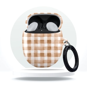 Puede incluir: Funda para auriculares con estampado de cuadros marrones y blancos, con un anillo negro. La funda está abierta, revelando auriculares blancos. La funda está sobre un pedestal blanco sobre un fondo blanco.