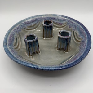 May include: A handmade ceramic candle holder with three cylindrical holders. The holder is a round dish with a blue and purple rim and a gray interior. The candle holders are blue and gray.