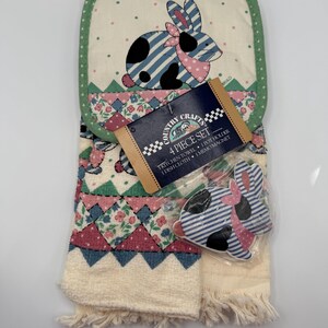 May include: A Country Crafts 4-piece kitchen set featuring a bunny design. The set includes a kitchen towel, pot holder, dish cloth, and a memo magnet. The bunny has blue stripes, black spots, and a pink bow. The towel has a cream fringe.