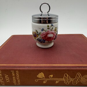 May include: A white porcelain egg coddler featuring a colorful floral pattern with pink roses and blue flowers. The container has a polished silver-tone metal lid with a circular ring handle on top, resting on a pedestal base with a smooth finish.