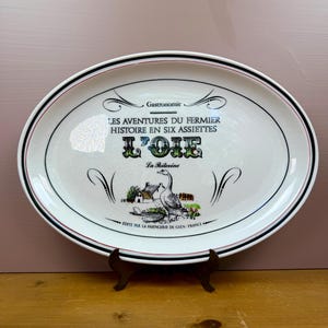 May include: An oval white ceramic platter featuring a black and red rim. The center displays the text "LES AVENTURES DU FERMIER HISTOIRE EN SIX ASSIETTES L'OIE" with a graphic of a goose and a farm, marked "EDITE PAR LA FAIENCERIE DE GIEN FRANCE".