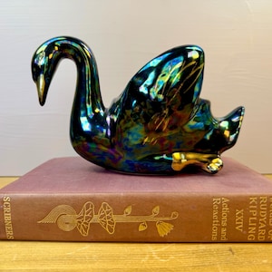 May include: An iridescent dark green and blue ceramic swan figurine with gold accents on the beak and feet, resting on a brown hardcover book titled The Works of Rudyard Kipling, Actions and Reactions, XXIV, published by Scribners.