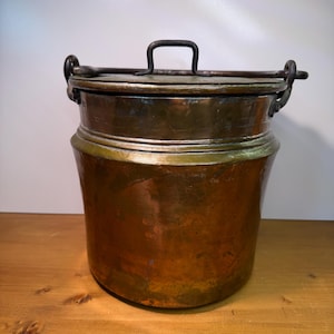 May include: A copper-colored metal pot with a lid and a handle. The pot has a cylindrical shape with a slightly flared base and a dark handle on top. The lid is attached with hinges. The pot is sitting on a wooden surface.