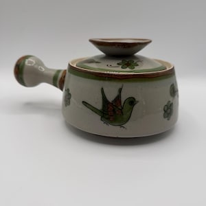 May include: A ceramic soup bowl with a lid, featuring a handle and a bird design. The bowl is primarily light gray with green and brown accents. The bird is green with red and orange details. The lid has a small knob.