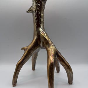 May include: A bronze-colored, tree-shaped candle holder. The candle holder has a textured surface and a flared base with three legs. The top has a small, circular opening for a candle. The overall design is organic and artistic.