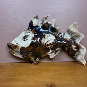 May include: A ceramic wall sculpture shaped like a fish with a glossy finish. The piece features a marbled color palette of cream, dark brown, and black, with textured scales and fins. The fish is depicted in a side profile with a prominent eye.