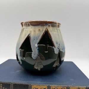 May include: A ceramic candle holder with a dark green base and a light blue and brown top. The holder has cut-out shapes of trees and a castle. The candle holder is sitting on a blue book.