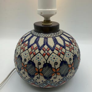 May include: A decorative ceramic table lamp with a round base. The base features a cream-colored background with blue, orange, and black geometric patterns. The lamp has a white bulb socket and a brown base.