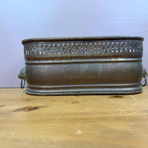 May include: An oval-shaped metal planter with a weathered bronze finish. The upper rim features a decorative openwork lattice pattern. Each side is adorned with a sculpted lion head handle that holds a small, thin metal ring for easy carrying.