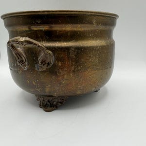 May include: A round, metallic brass pot with a weathered, tarnished patina. The vessel features two ornate, curved side handles and rests on three decorative, scrolled feet. The surface shows visible signs of age, oxidation, and discoloration.