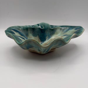 May include: A shallow ceramic bowl with a wavy, ruffled rim. The glaze features a blended pattern of deep teal, light blue, and creamy white tones. The interior surface is smooth and glossy, while the exterior base shows a natural clay finish.