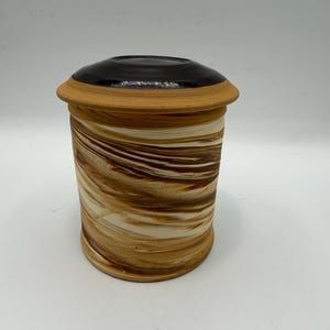 May include: A cylindrical ceramic jar with a lid. The jar features a swirling pattern of brown, tan, and cream colors. The lid is a dark brown color with a tan rim. The jar is likely handmade and suitable for storage.