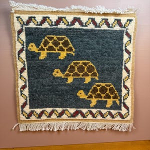 May include: A square woven wall hanging featuring a dark blue center panel with three brown and yellow turtles. The border consists of a white background with a repeating red and yellow zigzag pattern. The piece has white fringe on the top and bottom.