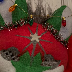 May include: A festive Christmas mask made from red, green, and white felt. The mask features a star design and is decorated with a string of colourful Christmas lights and tinsel. The mask has a mesh area for the eyes.