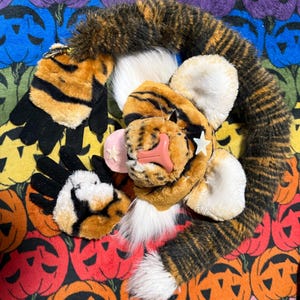 May include: A plush tiger tail and paw set with black stripes on orange fur. The tiger head has white fur, pink nose, and a white star. The background is a colourful Halloween-themed pattern with jack-o'-lanterns.