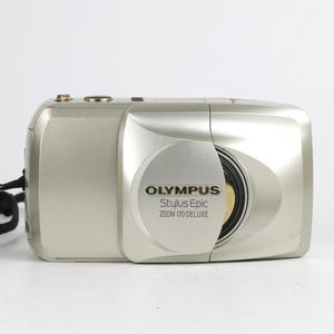 May include: A silver Olympus Stylus Epic Zoom 170 Deluxe point-and-shoot camera with a sliding lens cover. The front features the brand name and model text in black. A black wrist strap is attached to the left side of the compact camera body.