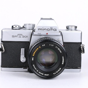 以下が含まれることがあります： 中央にMC Rokkor-X PF 50mm f/1.7レンズを搭載した、シルバーとブラックのミノルタSRT 201 35mmフィルムカメラ。カメラ本体にはテクスチャ加工された黒いグリップ、ダイヤル付きのシルバーのトッププレートがあり、前面のプリズムハウジングにはミノルタのロゴが印字されています。