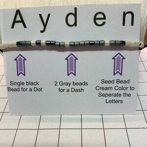 May include: A close-up of a white card with the word "Ayden" written in black letters. The card shows a string of beads with black, gray, and white beads. The card also includes instructions for using the beads to create the name "Ayden".