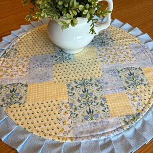 May include: A round, patchwork placemat with a ruffled edge. The placemat features a mix of floral and gingham patterns in yellow and blue. A white ceramic vase with greenery sits on top.