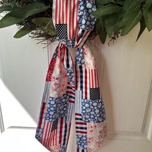 May include: A patriotic wreath decoration featuring a fabric scarf with a patchwork design. The scarf displays American flag motifs, including stars and stripes in red, white, and blue. The wreath has green leaves and brown twigs.