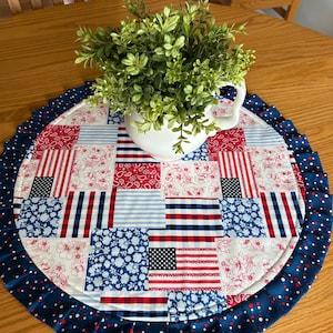 May include: A round patriotic placemat with a patchwork design featuring red, white, and blue patterns, including floral prints, stripes, and American flags. A white pitcher filled with greenery sits in the center. The placemat has a ruffled edge.