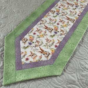 May include: A decorative table runner featuring a repeating pattern of bunnies and flowers on a white background. The runner is bordered by a light purple fabric and a speckled green fabric. The runner is placed on a gray quilted surface.
