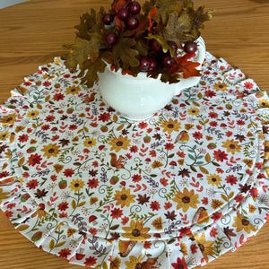 May include: A round fabric placemat featuring a white background with a colorful autumn-themed pattern of sunflowers, red flowers, brown acorns, mushrooms, squirrels, and birds, finished with a pleated ruffled edge and a white ceramic pitcher.