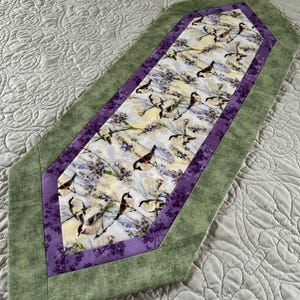 May include: A decorative table runner featuring a central panel with a bird and floral print, framed by a purple border and a green outer edge. The runner is designed with a pointed end on each side.