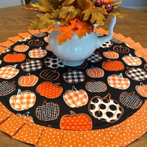 May include: A white ceramic pitcher with a curved handle holds fall-themed foliage. The pitcher sits on a round table runner with a black background and orange pumpkin patterns. The runner has a pleated orange border with white polka dots.