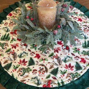 May include: A round Christmas-themed table topper with a cream-colored center featuring hedgehogs, red berries, and green trees. A green pleated border surrounds the center. A glass candle holder with a brown candle and a green and red wreath sits in the center.