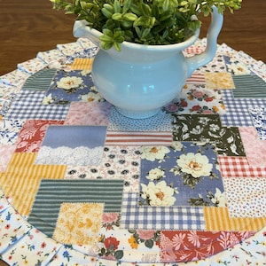May include: A white ceramic pitcher with a curved handle holds green foliage, set on a patchwork placemat. The placemat features a variety of floral and patterned fabrics in shades of blue, pink, yellow, and white, with a ruffled edge.