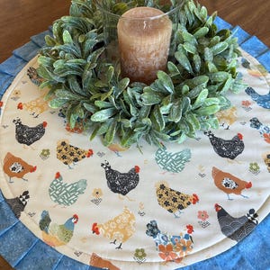 May include: A decorative centerpiece featuring a beige candle surrounded by a green wreath, set on a round placemat. The placemat has a blue ruffled edge and a pattern of colorful chickens.