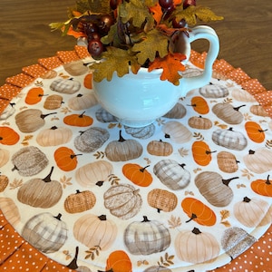 May include: A white ceramic mug filled with fall foliage sits on a round placemat. The placemat features a pumpkin pattern in shades of orange, brown, and cream, with a ruffled orange polka dot border.