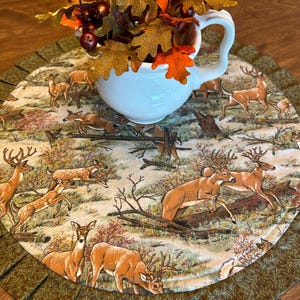 May include: A white ceramic jug filled with autumn leaves sits on a round table runner. The runner features a deer scene with a green pleated border. The deer are brown and white, set against a backdrop of trees and foliage.