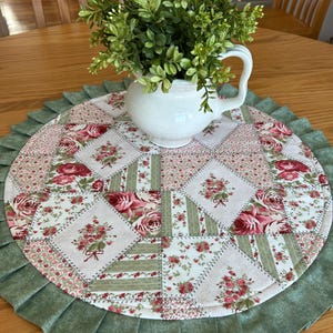 May include: A white ceramic jug with a handle contains a green plant arrangement, placed on a round, floral-patterned placemat. The placemat has a patchwork design with red roses and a green ruffled edge, on a wooden table.