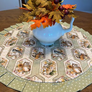 May include: A white ceramic pitcher holds a fall arrangement of orange and yellow leaves and berries. The pitcher sits on a round placemat with a light green border and a repeating pattern of woodland creatures and sunflowers.
